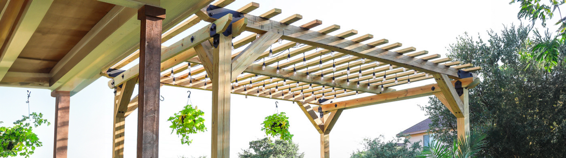 A wooden pergola structure with sturdy beams and slats, situated outdoors with hanging plants and greenery in the background.