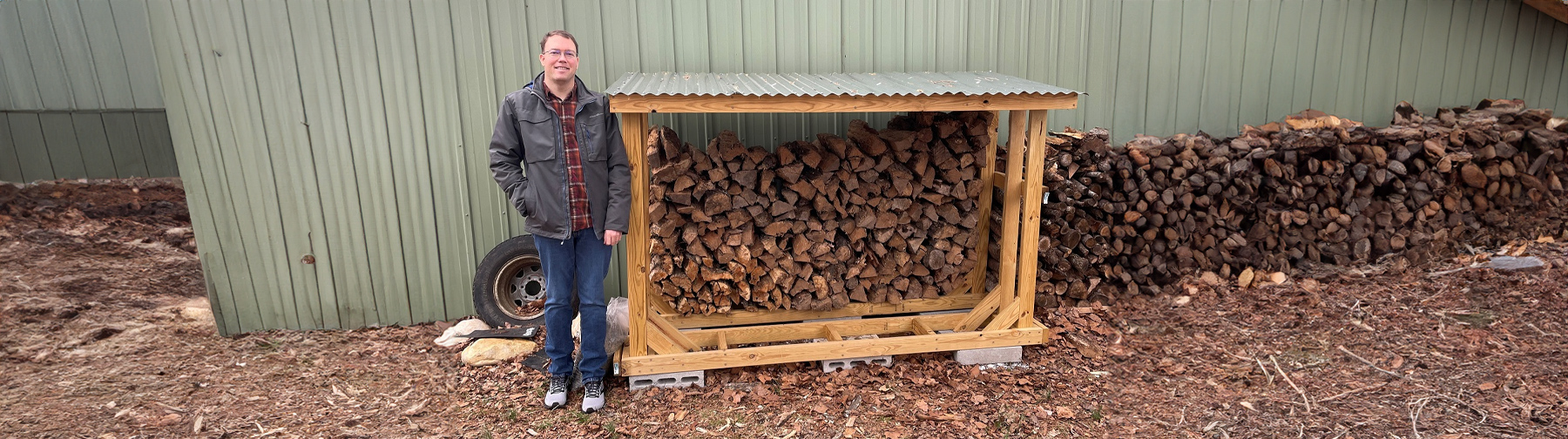 A man standing next to a neatly stacked firewood shelter made of wood, featuring a corrugated metal roof, with a large pile of firewood in the background.