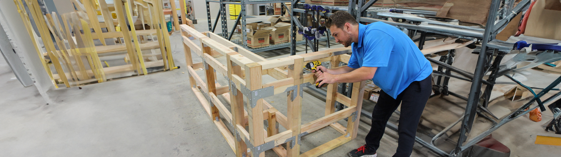 A worker in a blue shirt using a power drill to assemble a wooden framework structure in an industrial workshop setting.