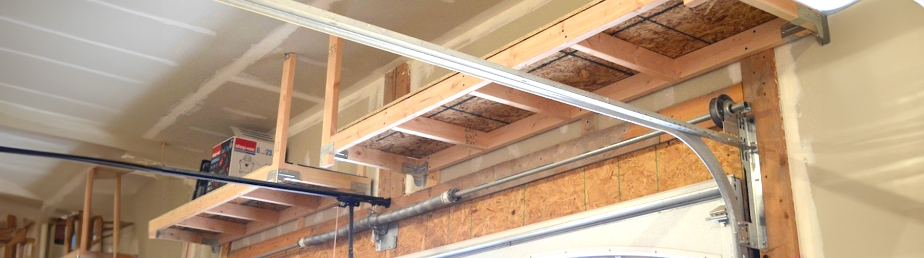 A wooden overhead storage shelf mounted in a garage, supported by beams and brackets, with a garage door track running below it.