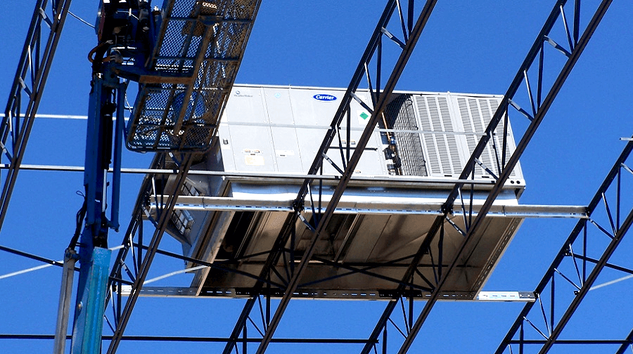 A rooftop HVAC unit being installed on a steel support frame using a lift, with a clear blue sky in the background.