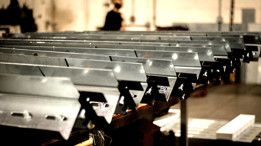 A row of galvanized steel framing components lined up on a workbench in a manufacturing facility, with a worker visible in the background.